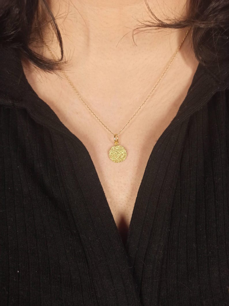 May include: A gold-colored pendant necklace with a circular charm. The charm has an intricate design and is suspended from a delicate chain. The necklace is worn against a black top.