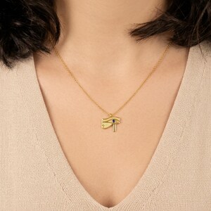 May include: A gold-colored pendant necklace featuring the Eye of Horus symbol. The pendant has a small blue gemstone and hangs from a delicate gold chain. The necklace is displayed on a person wearing a beige top.