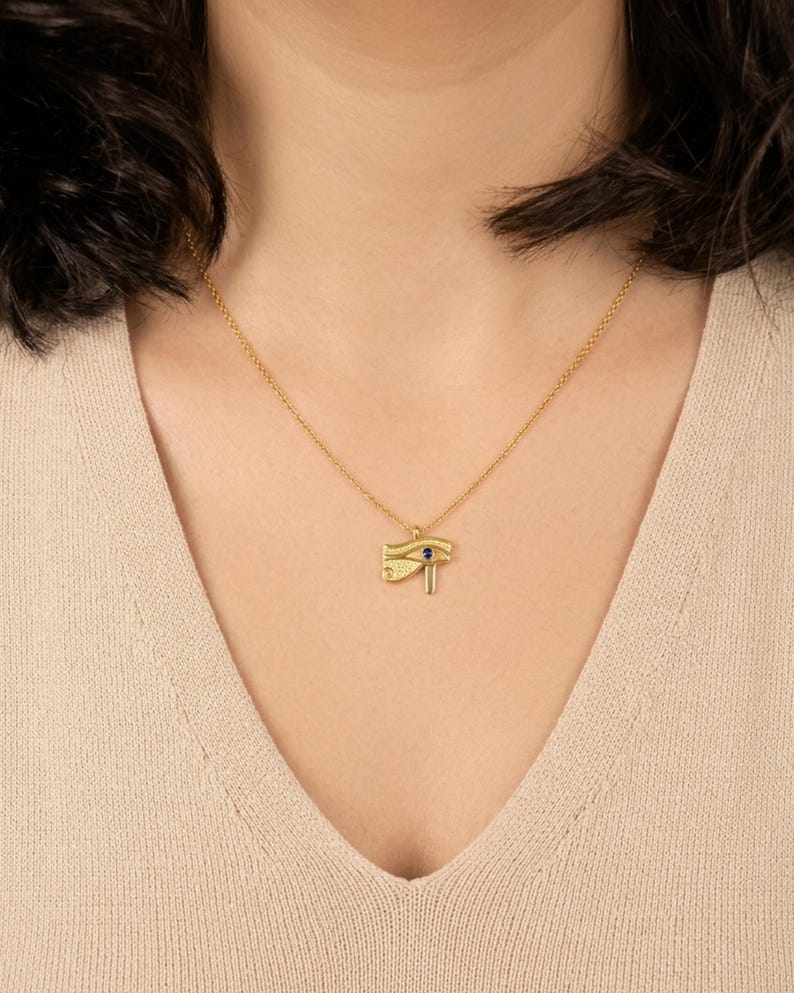 May include: A gold-colored pendant necklace featuring the Eye of Horus symbol. The pendant has a small blue gemstone and hangs from a delicate gold chain. The necklace is displayed on a person wearing a beige top.