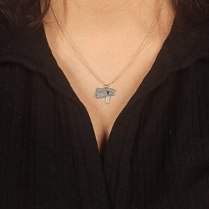 May include: A silver-toned necklace featuring a pendant in the shape of the Eye of Horus. The pendant has intricate detailing with a blue gemstone accent. The necklace is displayed against a black top.