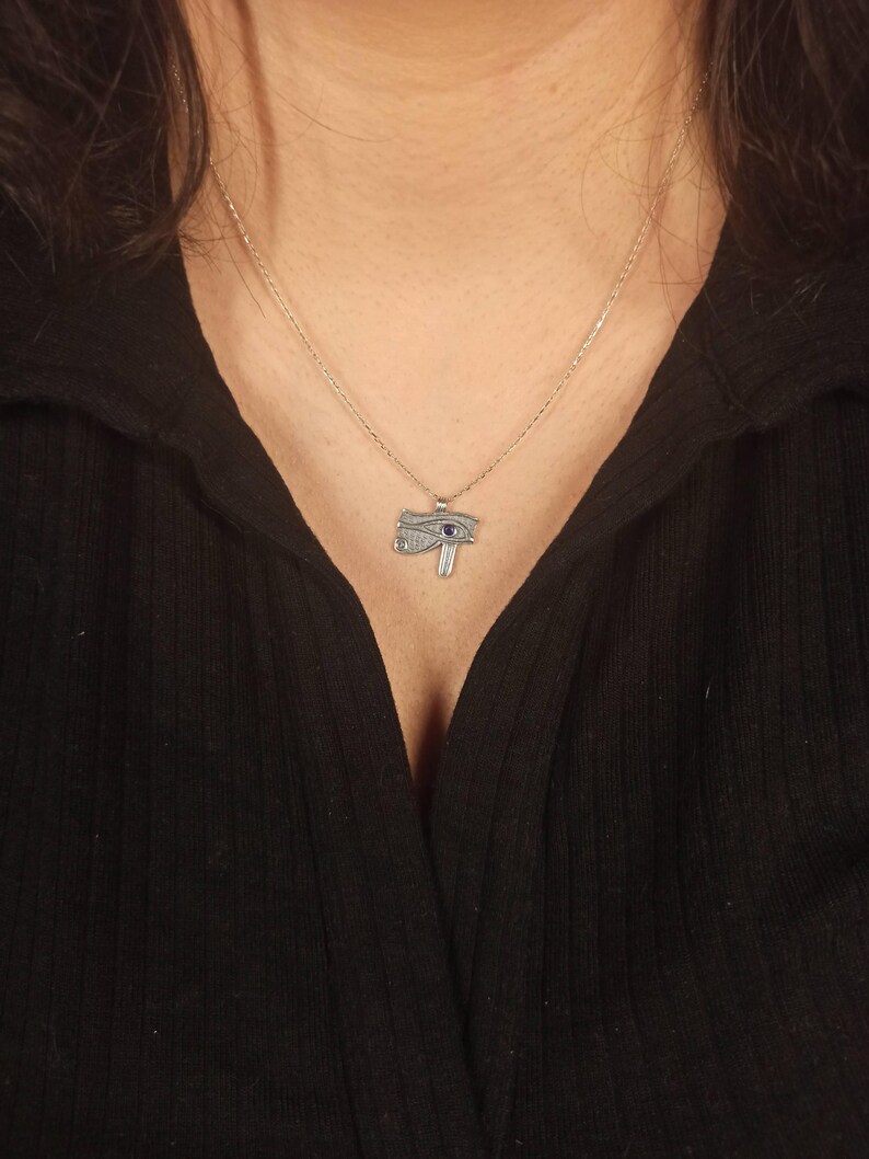 May include: A silver-toned necklace featuring a pendant in the shape of the Eye of Horus. The pendant has intricate detailing with a blue gemstone accent. The necklace is displayed against a black top.