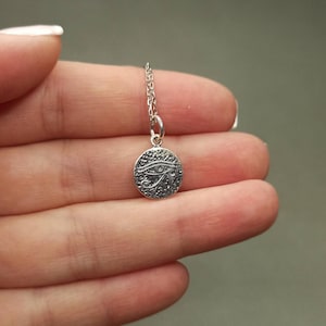 May include: A silver-colored pendant necklace featuring a small, round charm. The charm has a textured surface and an engraved Eye of Horus design. The necklace chain is delicate and silver. The pendant is held between fingers.