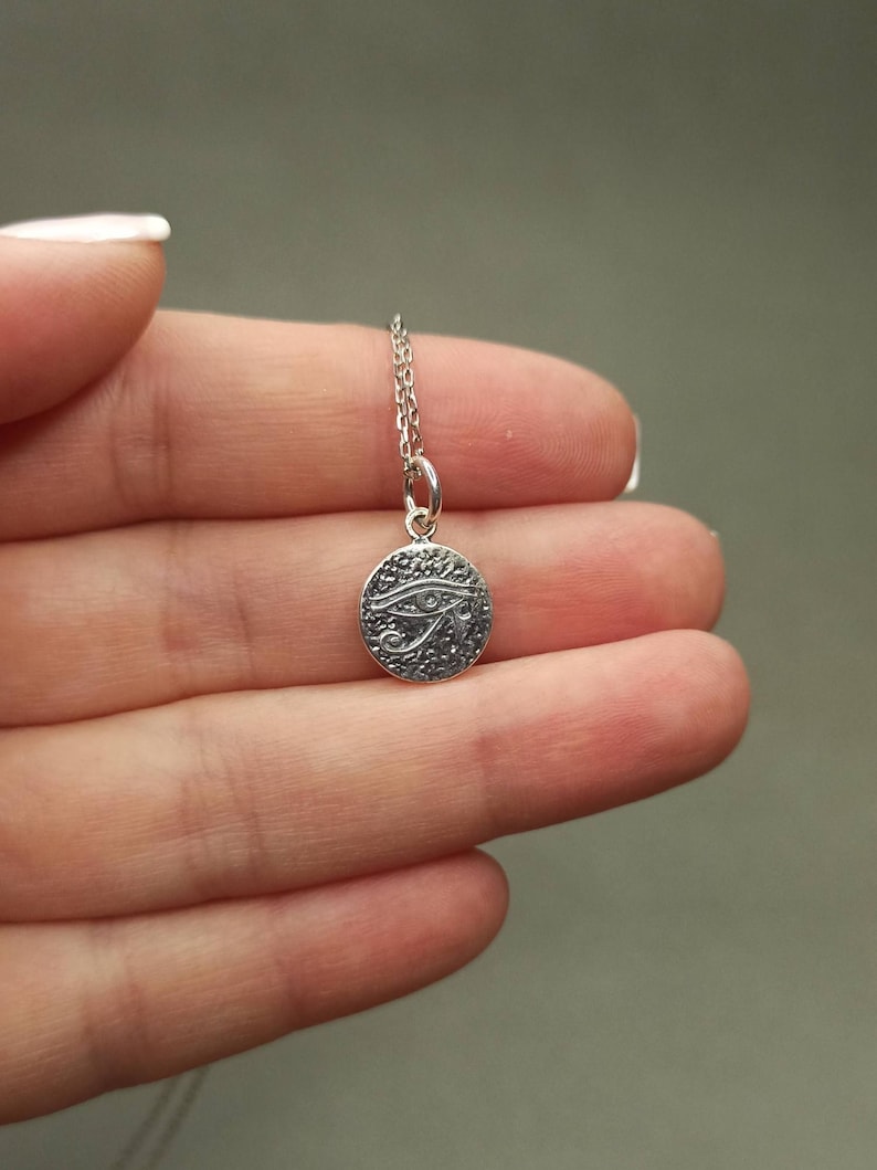 May include: A silver-colored pendant necklace featuring a small, round charm. The charm has a textured surface and an engraved Eye of Horus design. The necklace chain is delicate and silver. The pendant is held between fingers.