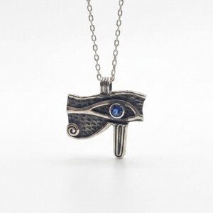 May include: A silver-toned pendant necklace featuring the Eye of Horus design. The pendant has a textured surface with a blue gemstone in the center of the eye. The necklace chain is a delicate silver color.