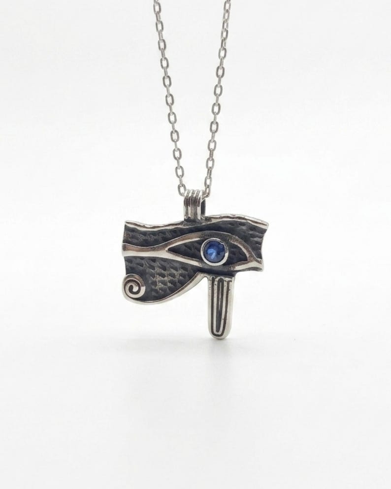 May include: A silver-toned pendant necklace featuring the Eye of Horus design. The pendant has a textured surface with a blue gemstone in the center of the eye. The necklace chain is a delicate silver color.
