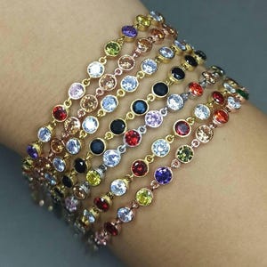 May include: A set of seven bracelets with colorful gemstones and clear rhinestones. Each bracelet features a different color gemstone, including red, orange, yellow, green, blue, purple, and black.
