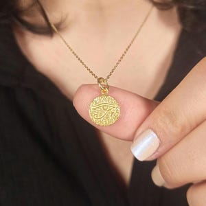 May include: A gold-colored pendant necklace featuring a circular charm with an engraved Eye of Horus design. The charm is suspended from a delicate gold chain. The necklace is shown against a dark background.