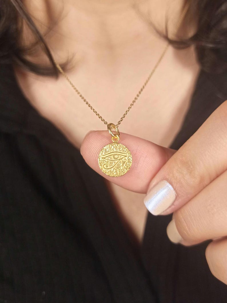 May include: A gold-colored pendant necklace featuring a circular charm with an engraved Eye of Horus design. The charm is suspended from a delicate gold chain. The necklace is shown against a dark background.