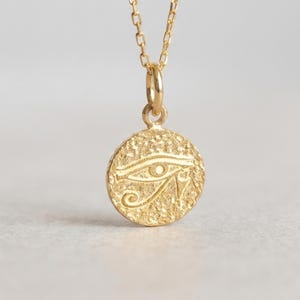 May include: A gold-colored pendant necklace featuring a circular charm with an engraved Eye of Horus symbol. The charm is attached to a delicate chain. The necklace is displayed against a neutral background, highlighting the jewelry's design.