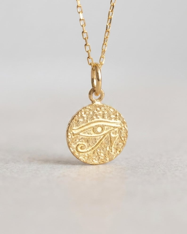May include: A gold-colored pendant necklace featuring a circular charm with an engraved Eye of Horus symbol. The charm is attached to a delicate chain. The necklace is displayed against a neutral background, highlighting the jewelry's design.
