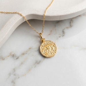 May include: A gold-colored necklace featuring a circular pendant with an embossed Eye of Horus design. The pendant hangs from a delicate chain. The necklace is displayed on a white marble surface.