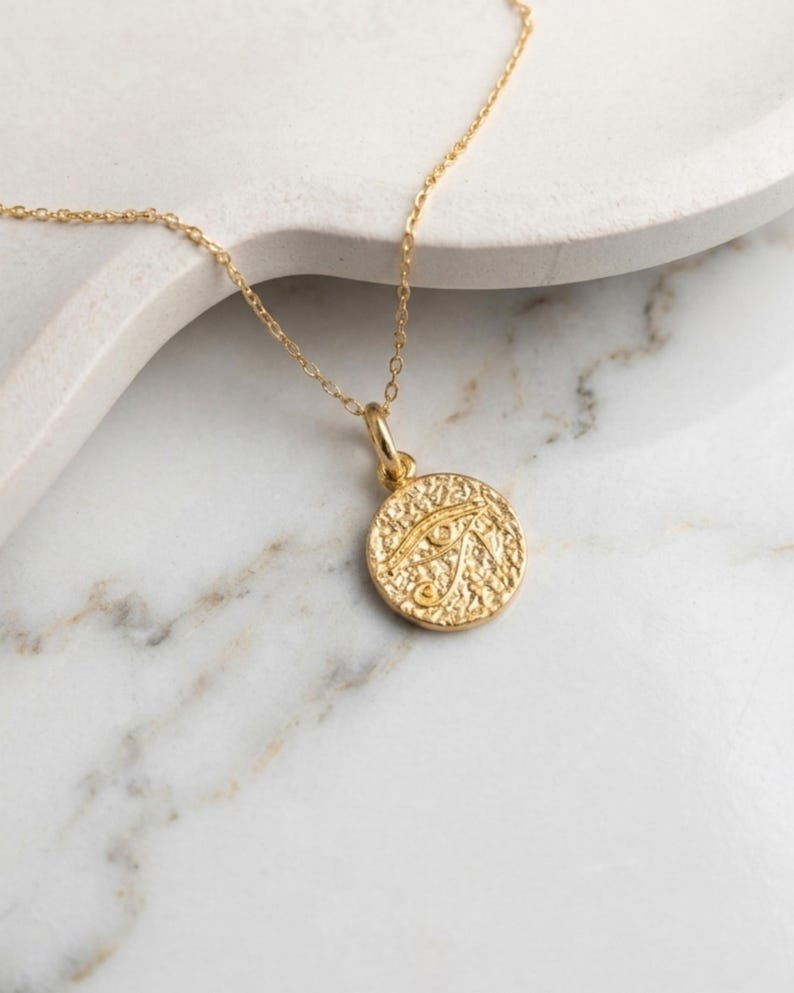 May include: A gold-colored necklace featuring a circular pendant with an embossed Eye of Horus design. The pendant hangs from a delicate chain. The necklace is displayed on a white marble surface.