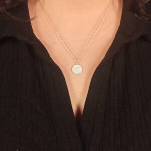 May include: A silver necklace with a small, round pendant. The pendant features a detailed design, possibly an eye symbol, and hangs from a delicate chain. The necklace is worn against a black top.