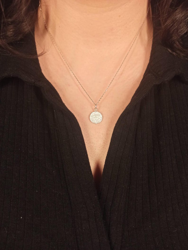 May include: A silver necklace with a small, round pendant. The pendant features a detailed design, possibly an eye symbol, and hangs from a delicate chain. The necklace is worn against a black top.
