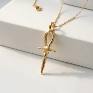 Puede incluir: Un collar con colgante Ankh dorado. El colgante, una cruz estilizada con un lazo, cuelga de una delicada cadena. El collar tiene un acabado texturizado y se presenta sobre una superficie blanca. El Ankh es un símbolo de vida.