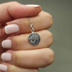 May include: A silver-colored pendant necklace featuring a circular charm with an engraved Eye of Horus design. The charm is suspended from a delicate chain. The necklace is held up against a neutral background.