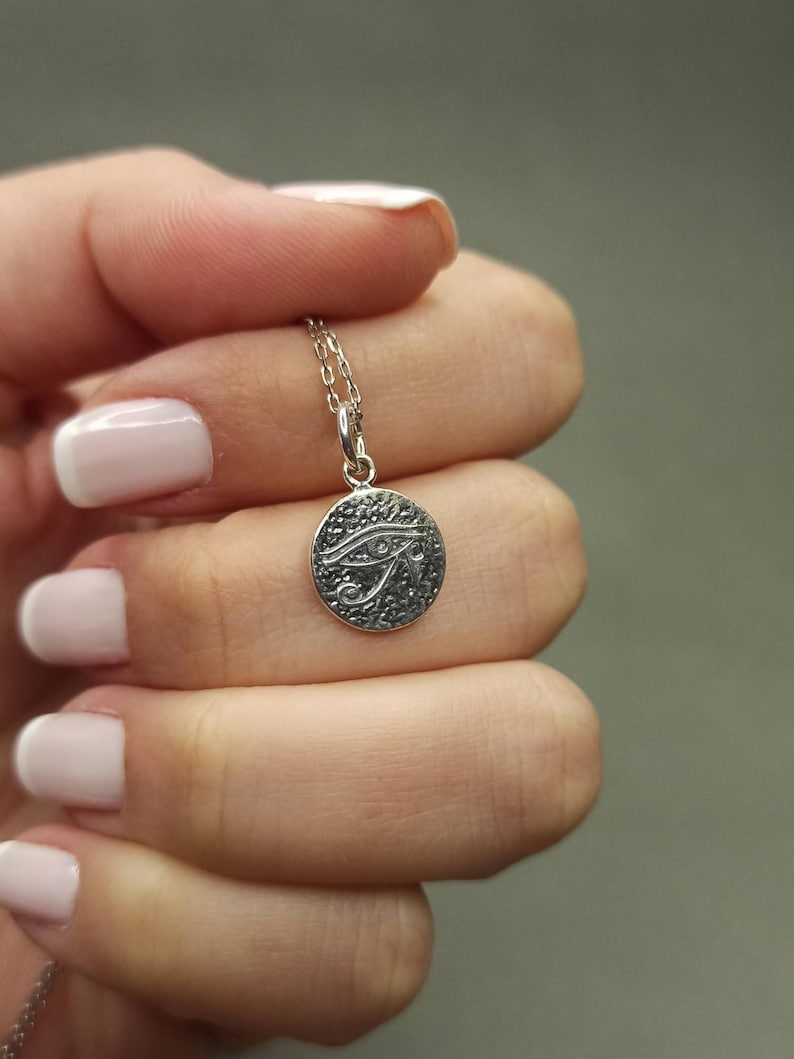 May include: A silver-colored pendant necklace featuring a circular charm with an engraved Eye of Horus design. The charm is suspended from a delicate chain. The necklace is held up against a neutral background.