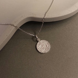 May include: A silver necklace featuring a circular pendant with an eye design. The pendant has a textured surface and is suspended from a delicate chain. The necklace is displayed on a neutral background, highlighting its simple elegance.