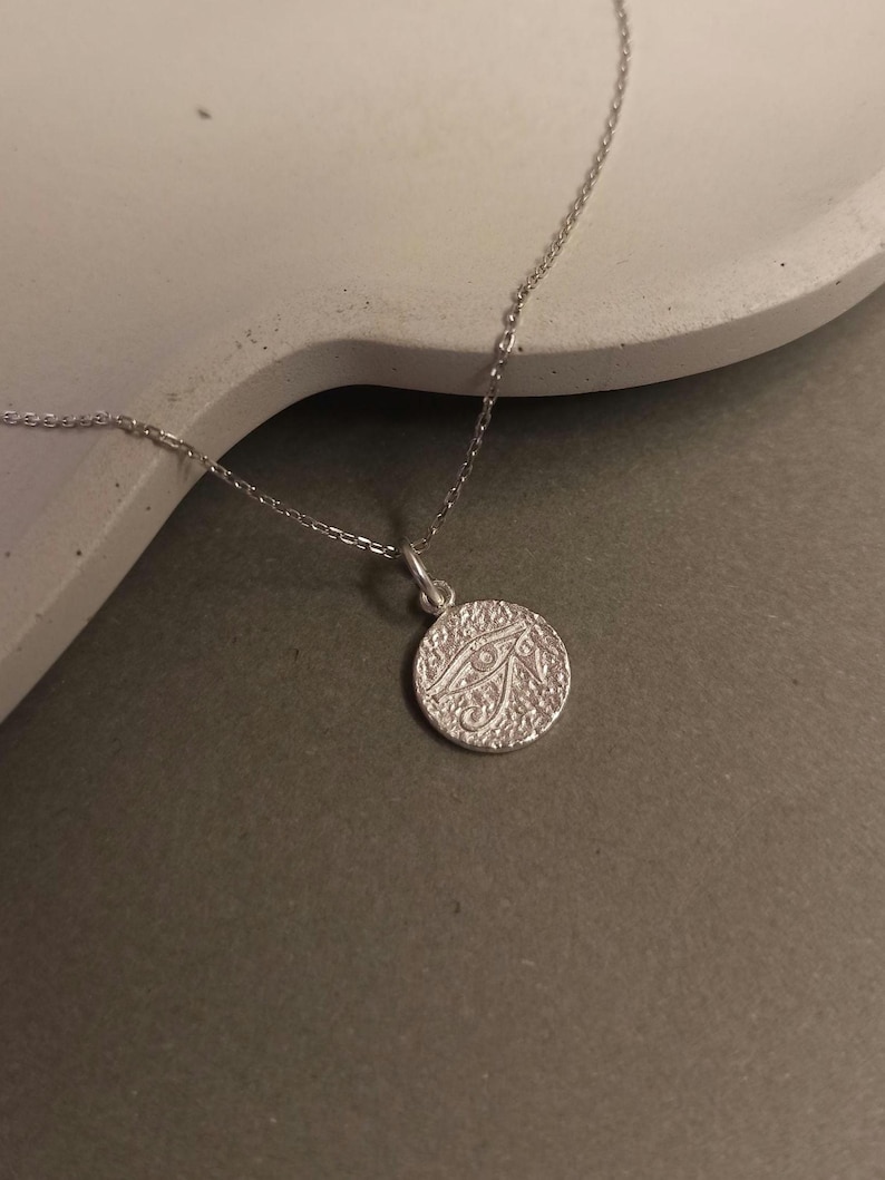 May include: A silver necklace featuring a circular pendant with an eye design. The pendant has a textured surface and is suspended from a delicate chain. The necklace is displayed on a neutral background, highlighting its simple elegance.