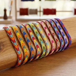 May include: A collection of colorful woven bracelets displayed on a wooden dowel. The bracelets feature a variety of rainbow hues, including red, orange, yellow, green, blue, and purple. The bracelets are approximately 0.5 inches wide.