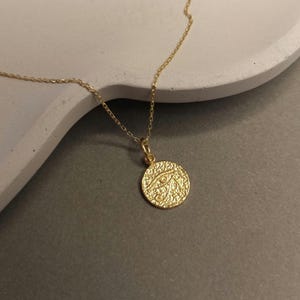 May include: A gold-colored necklace with a circular pendant. The pendant features a textured surface and an eye design. The necklace is displayed on a light gray surface.