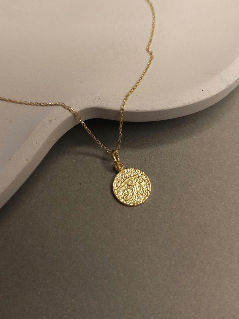 May include: A gold-colored necklace with a circular pendant. The pendant features a textured surface and an eye design. The necklace is displayed on a light gray surface.