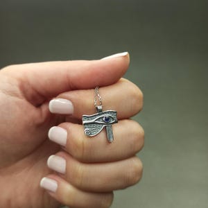 May include: A silver-toned pendant necklace featuring the Eye of Horus design. The pendant has a blue gemstone accent and is suspended from a delicate chain. The necklace is held in a hand against a gray background.