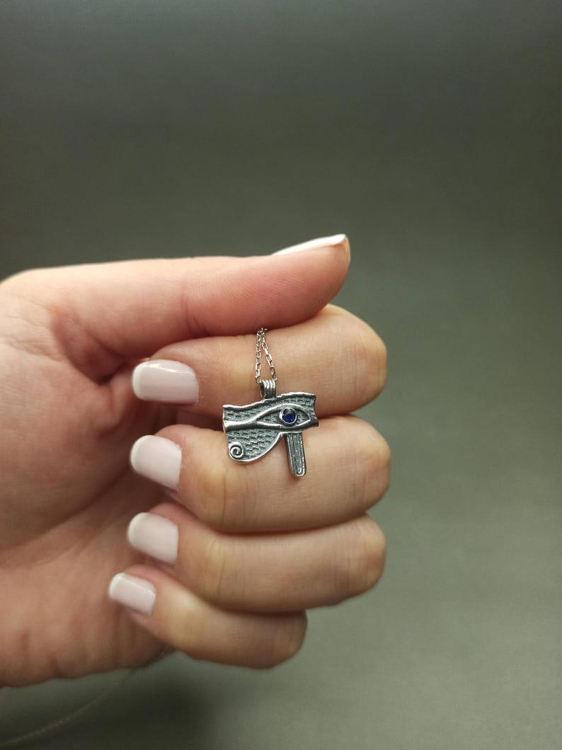 May include: A silver-toned pendant necklace featuring the Eye of Horus design. The pendant has a blue gemstone accent and is suspended from a delicate chain. The necklace is held in a hand against a gray background.