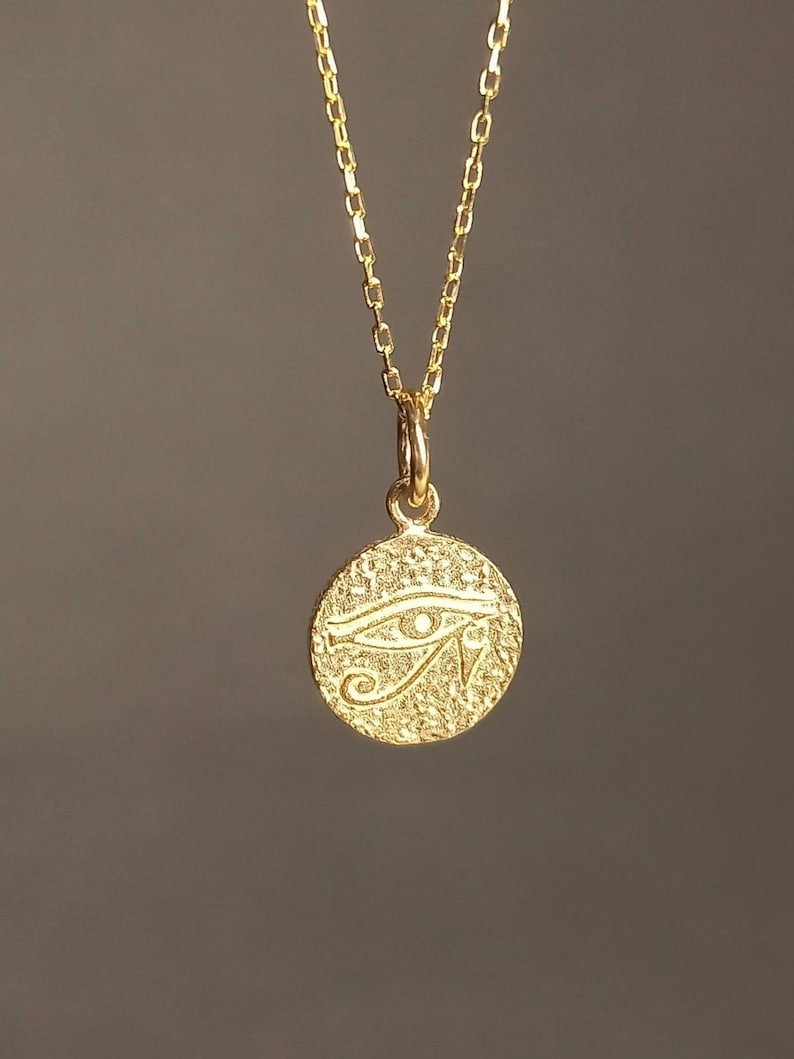 May include: A gold-colored necklace featuring a circular pendant with a textured surface. The pendant displays the Eye of Horus symbol, a stylized eye with intricate details. The chain is delicate and complements the pendant's design.