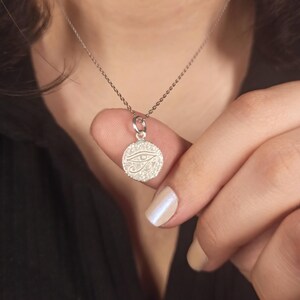 May include: A silver necklace featuring a circular pendant with an engraved Eye of Horus design. The pendant is held by a person. The necklace chain is delicate and the pendant is small.