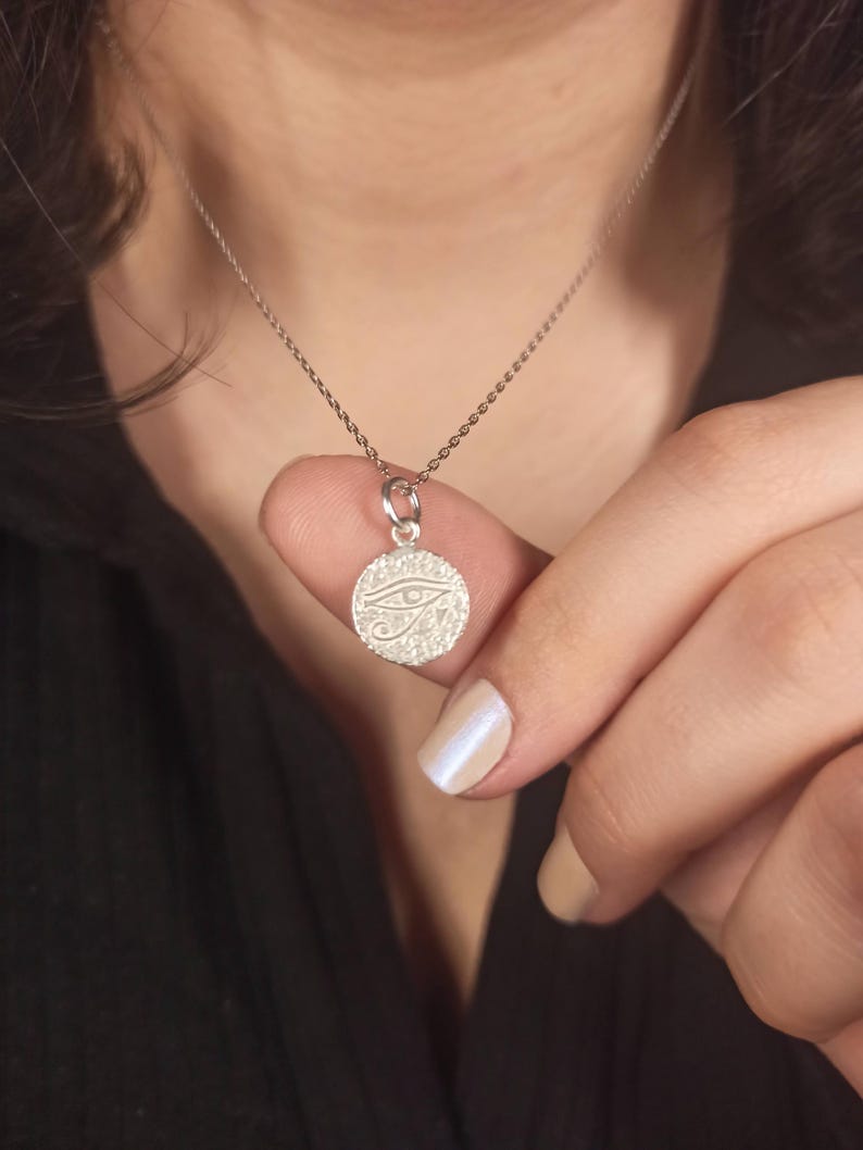 May include: A silver necklace featuring a circular pendant with an engraved Eye of Horus design. The pendant is held by a person. The necklace chain is delicate and the pendant is small.