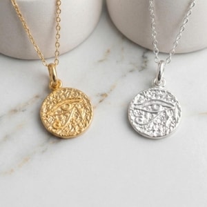 May include: Two necklaces with circular pendants featuring an eye design. One necklace is gold-toned, the other is silver-toned. Both pendants hang from delicate chains. The pendants have a textured surface and are displayed on a white marble surface.