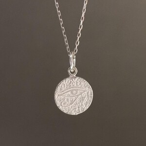 May include: A silver-colored necklace featuring a circular pendant with a textured surface. The pendant displays an engraved Eye of Horus symbol. The chain is delicate and hangs against a dark background, creating a simple, elegant design.
