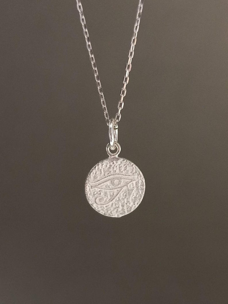 May include: A silver-colored necklace featuring a circular pendant with a textured surface. The pendant displays an engraved Eye of Horus symbol. The chain is delicate and hangs against a dark background, creating a simple, elegant design.