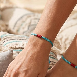 May include: A light blue braided cord bracelet and anklet set featuring a small, vibrant red wrapped accent detail. The jewelry pieces are constructed from thin, woven material and include adjustable cord ends for a secure fit on the wrist and ankle.