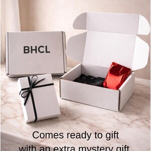 May include: White gift boxes with black ribbon and the letters "BHCL" printed on one. One box is open, revealing black tissue paper and a red package. The text "Comes ready to gift with an extra mystery gift" is at the bottom.
