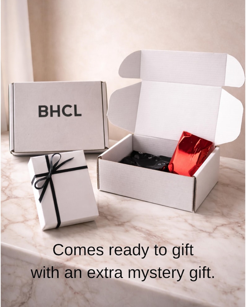 May include: White gift boxes with black ribbon and the letters "BHCL" printed on one. One box is open, revealing black tissue paper and a red package. The text "Comes ready to gift with an extra mystery gift" is at the bottom.