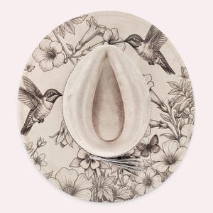 May include: A wide-brimmed beige hat featuring a detailed black line art illustration of two hummingbirds, several butterflies, and various floral patterns across the brim. The crown is indented and includes a small decorative tassel detail.