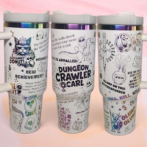 May include: Three white tumblers with iridescent metallic rims and handles, featuring a wrap-around graphic print of Dungeon Crawler Carl characters, skulls, swords, potions, and text including "Dungeon Crawler Carl" and "Goddammit Donut".
