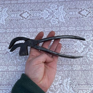 May include: A vintage, rusty, multi-tool pliers with a hammer head on one end and a claw on the other. The tool is held in a hand.
