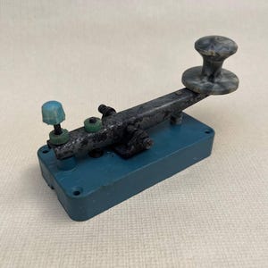 Peut inclure: Clé télégraphique vintage avec une base turquoise et un levier noir. La clé a un bouton gris marbré et un bouton bleu sur le côté gauche. Les composants métalliques montrent des signes d'usure. Un objet historique de communication.