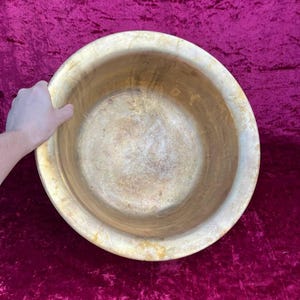 May include: A large, round wooden bowl with a light brown finish. The bowl has a wide rim and a deep interior. The bowl is held by a hand. The background is a deep red velvet.