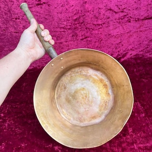 May include: A vintage brass pan with a long wooden handle. The pan has a wide, shallow bowl and is held against a deep pink background. The brass has a warm, aged patina, suggesting its use over time. The handle is attached with rivets.