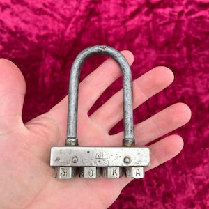 May include: A vintage metal combination lock with a silver finish. The lock features a U-shaped shackle and a series of lettered dials. The text "Я О К А Ш А" is visible on the dials, along with the text "ХМТ" and "U.1930".