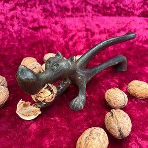 May include: A vintage, cast-iron nutcracker in the shape of a dog, with a walnut in its jaws. The dog-shaped nutcracker is black and sits on a red velvet surface, surrounded by walnuts. The nutcracker is approximately 6 inches long.