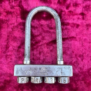 May include: A vintage combination padlock with a U-shaped shackle. The lock is made of metal with a weathered appearance. The combination dials display the numbers and letters. The lock is set against a pink background.