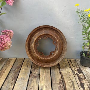 Könnte beinhalten: Eine kreisförmige, verwitterte Metallfelge mit einer dicken Schicht aus braunem Rost. Die Felge hat eine zentrale Öffnung, die von fünf gleichmäßig verteilten Bolzenlöchern umgeben ist. Die Metalloberfläche weist starke Oxidation auf.