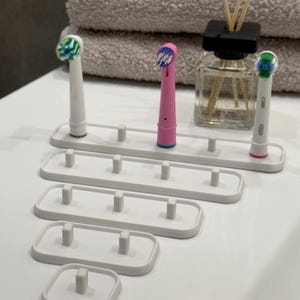 Könnte beinhalten: Ein Set weißer Zahnbürstenhalter in verschiedenen Größen, zur Aufnahme mehrerer elektrischer Zahnbürstenköpfe. Die Halter sind auf einer weißen Oberfläche angeordnet, mit drei Zahnbürsten. Im Hintergrund ein handtuch und ein Diffusor.