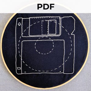 Peut inclure: Un cerceau de broderie noir avec une image brodée blanche d'une disquette. La disquette est soulignée de fil blanc et comporte une flèche blanche pointant vers le haut.