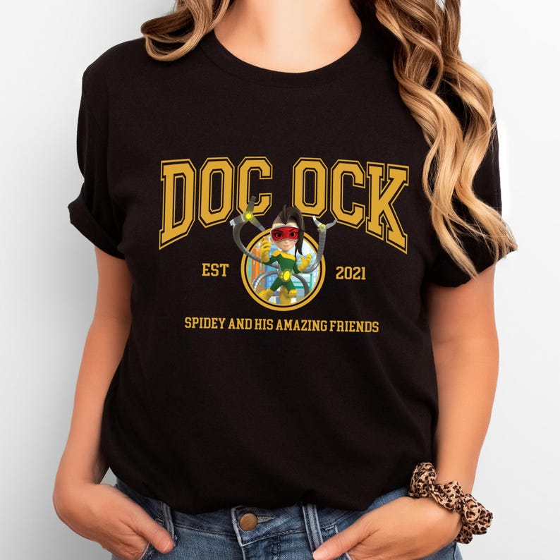 Pu&ograve; includere: T-shirt nero con design dorato "DOC OCK", "EST 2021" e un'illustrazione di un personaggio dei cartoni animati. Il testo "SPIDEY AND HIS AMAZING FRIENDS" &egrave; sotto. La maglietta &egrave; in materiale morbido.